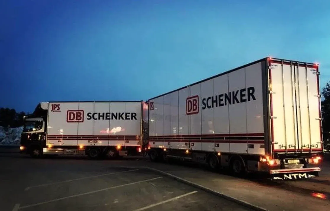 Två upplysta DB Schenker-ekipage uppställda på terminalplanen i mörker.