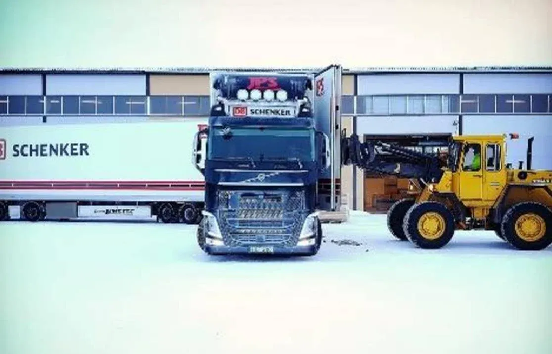 Blå Volvo med DB Schenker-skåp och en hjullastare framför verkstadsportarna.
