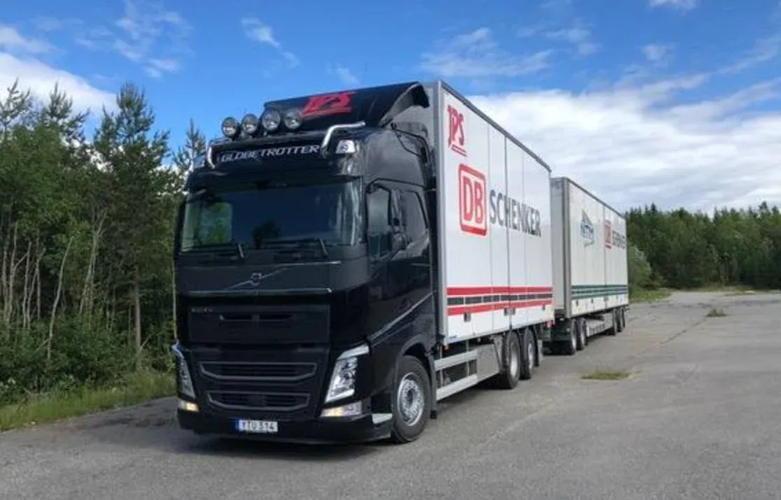 Svart Volvo dragbil med DB Schenker-skåp på grusplan omgiven av skog.
