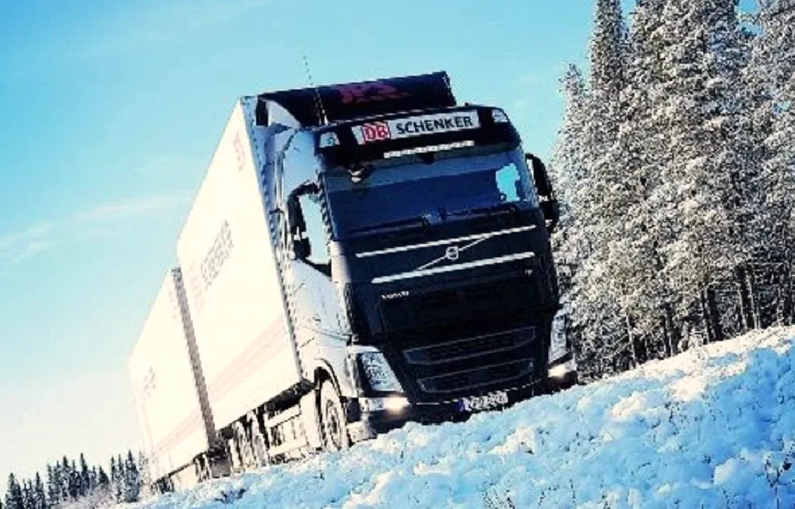 Volvo med DB Schenker-skåp på vinterväg genom snötyngd granskog.